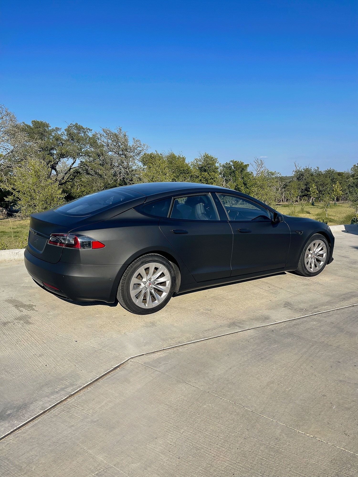 Black sedan parked on a sunlit concrete road beside trees under a clear blue sky