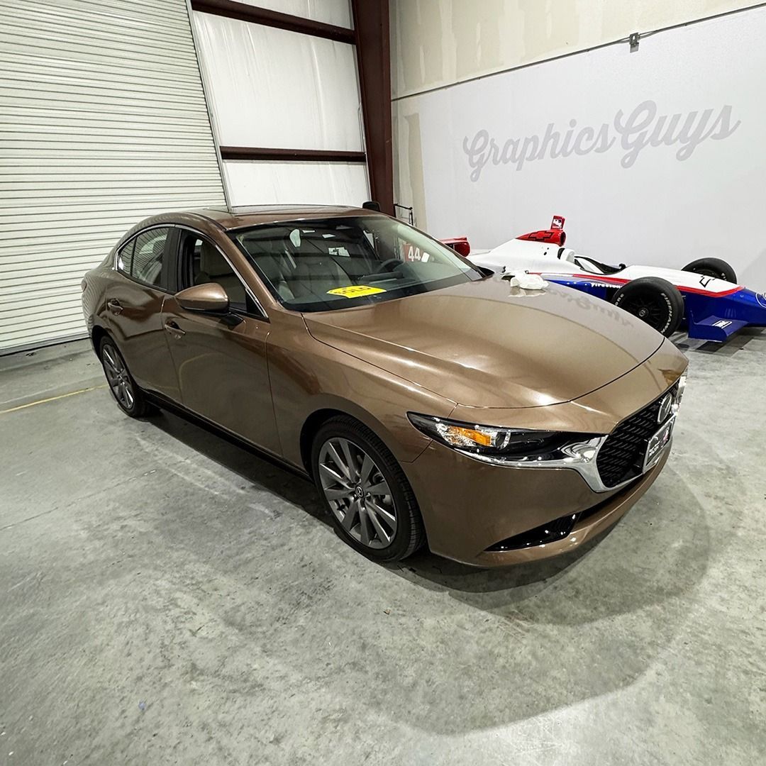 Brown Mazda hatchback in a garage beside a wall with “Graphics Guys” lettering and race cars.