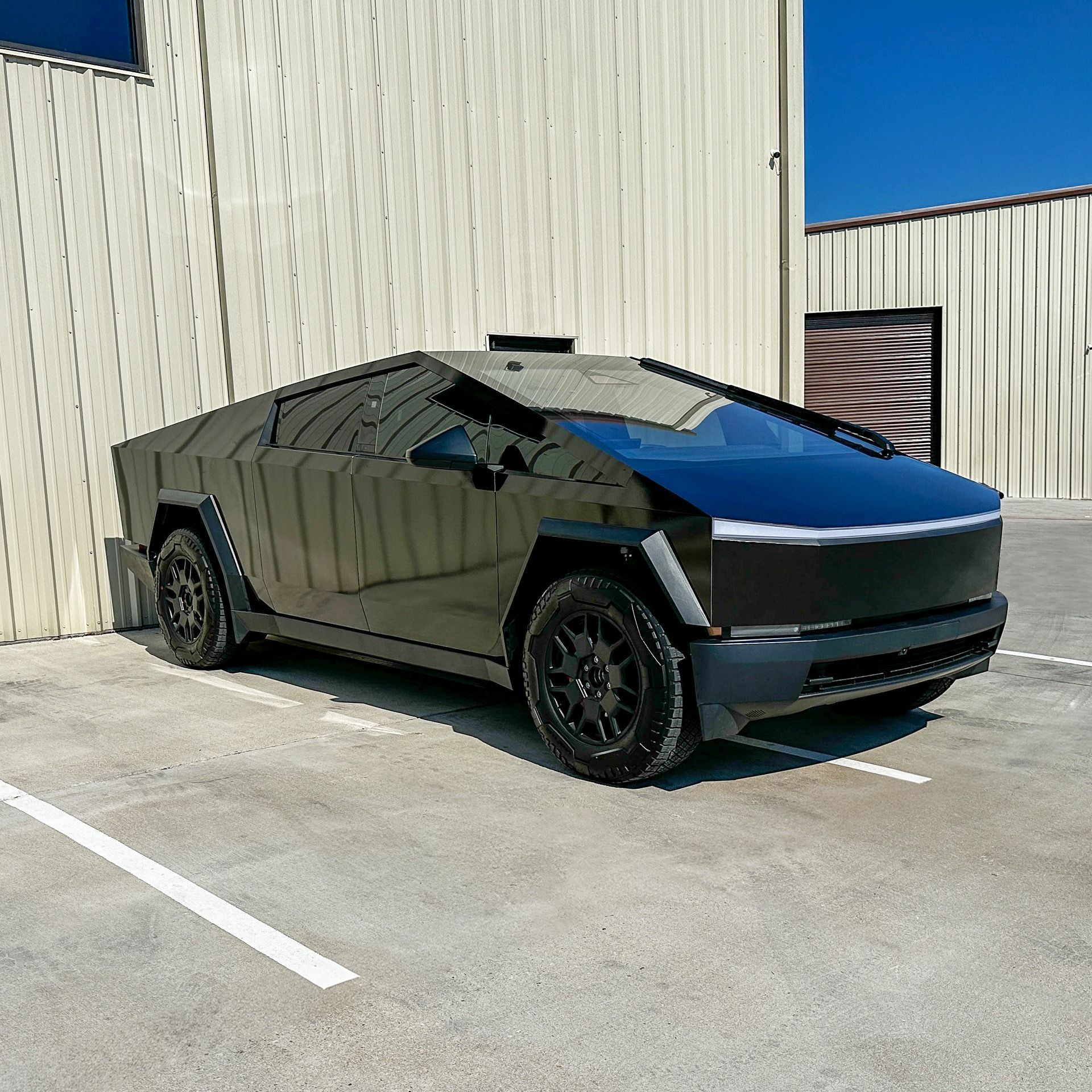 Matte black Tesla Cybertruck parked beside a beige industrial building on a sunny day