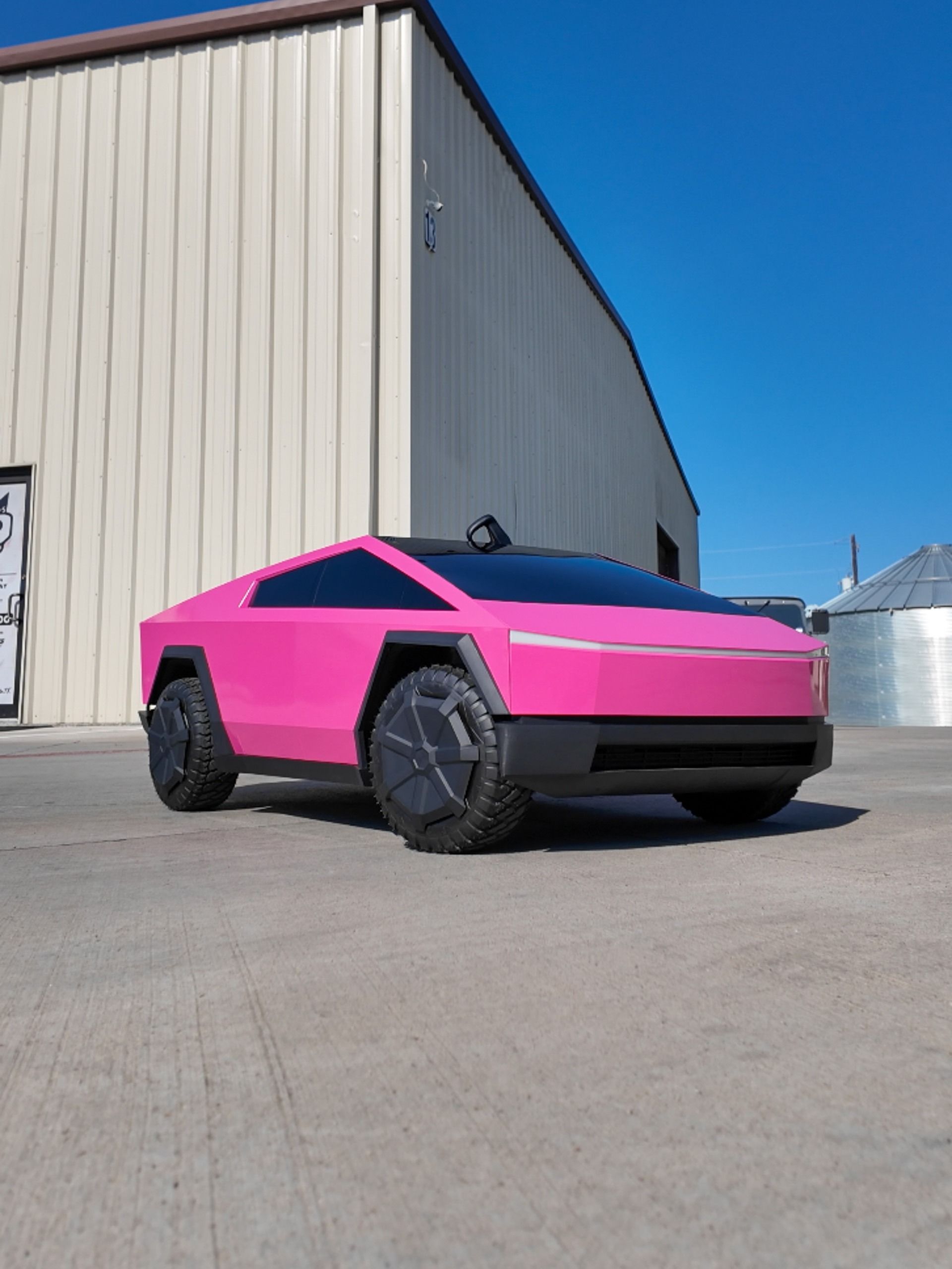 Bright pink angular Tesla Cybertruck parked on pavement beside a beige industrial building under a blue sky