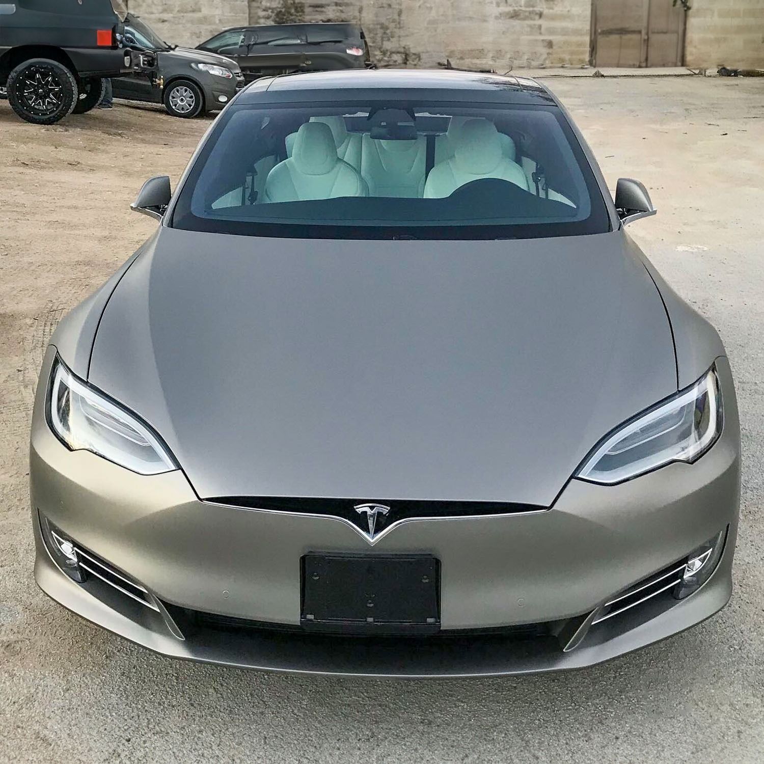 Front view of a matte gray Tesla sedan parked outdoors on a gravel lot.