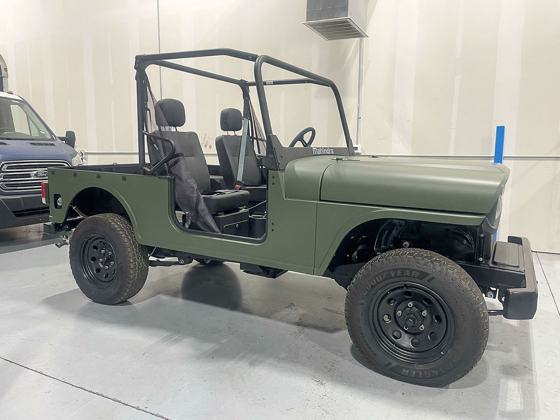 Olive green vintage off-road Jeep in a garage, viewed from the side with no doors and a roll cage