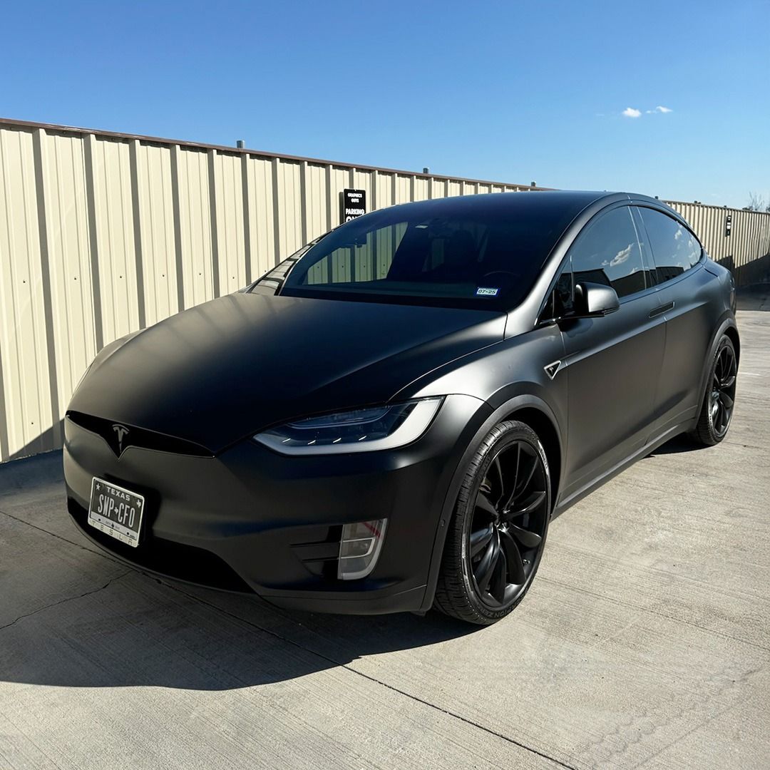 Black Tesla Model Y parked on a sunny street beside a beige fence