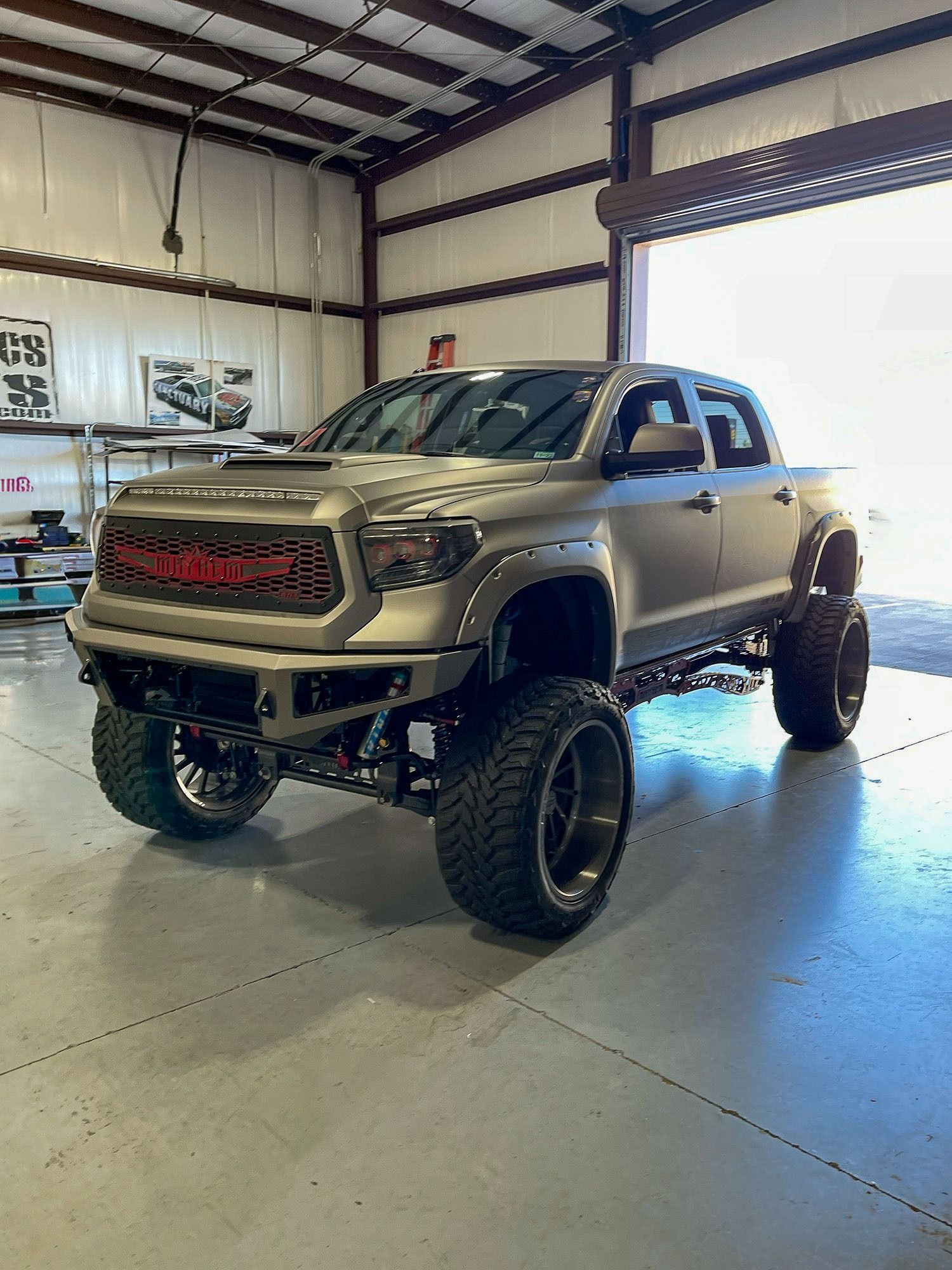 Lifted tan pickup truck in a garage with large off-road tires and red grille accents.