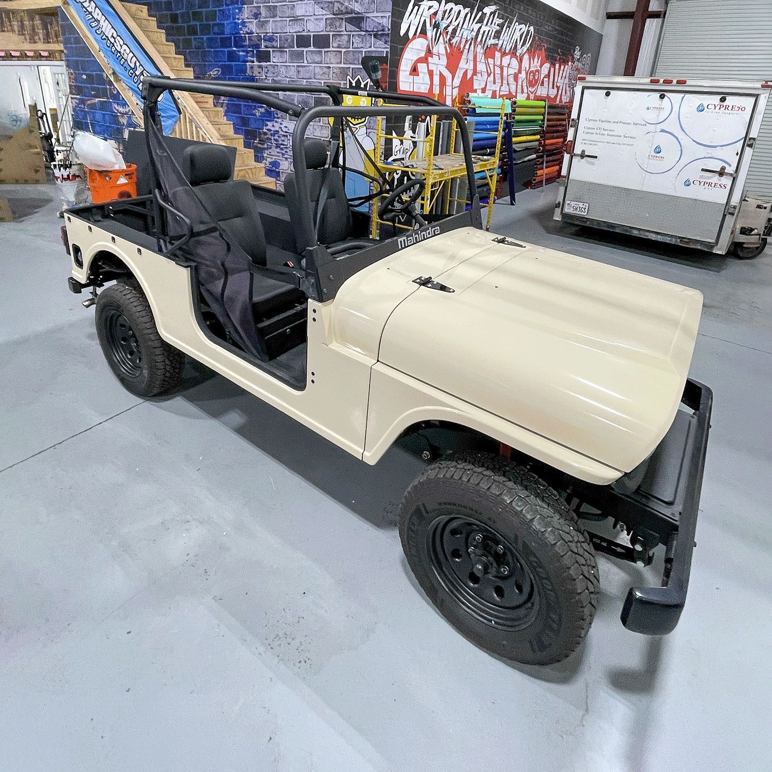 Beige open-top utility jeep with black seats, parked indoors near graffiti walls and equipment.