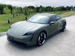 Matte green sports car parked on a concrete road beside a grassy field and trees.