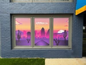 Storefront with three windows showing a pink-purple sunset cityscape and cacti mural.