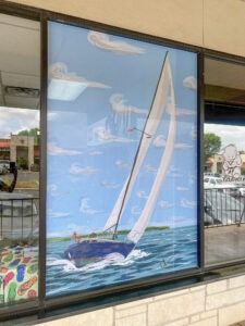 Sailboat mural in a window, with a white-sailed boat on blue water under a cloudy sky.