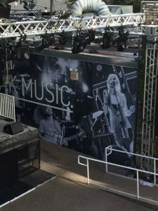 Outdoor concert stage with large black-and-white MUSIC banner and performers on a platform