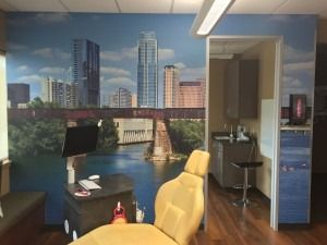 Office room with city mural, desk, monitor, beige chair, and doorway to a back room.