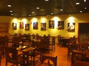 Warm-lit empty restaurant dining room with wooden tables, chairs, and wall art.