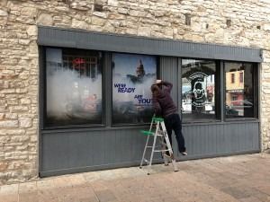 Person on a ladder applying graphics to a storefront window with stone facade.