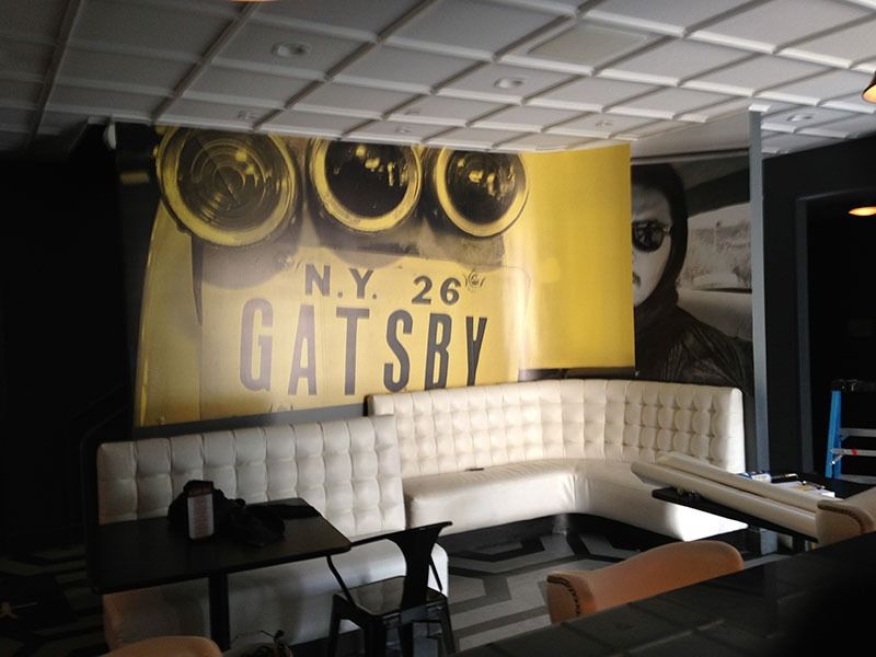 Dim lounge with white tufted seating and a yellow Gatsby wall mural under a grid ceiling.