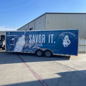 Blue truck trailer with “SAVOR IT.” and a large bottled drink graphic parked outside a warehouse.