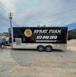 Black SPRAY FOAM trailer with phone number, parked on a gravel lot under a cloudy sky