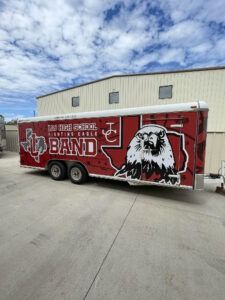 Red trailer with eagle and “BAND” graphics parked beside a building under a cloudy sky