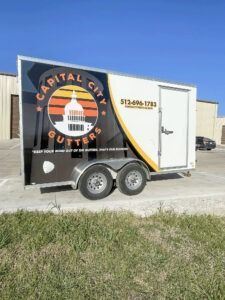 Black and white Capital City Outfitter trailer parked on gravel beside grass under blue sky