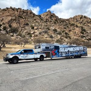 Blue pickup truck towing a GRAPHICS GUYS trailer on a road beside rocky desert hills