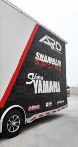 Black trailer with red stripe and racing sponsor logos, parked on a road