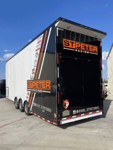 White STPETER Racing trailer with black rear door, parked outdoors on pavement under a blue sky