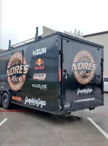Black race trailer with NORES logo and sponsor decals, parked in a lot outdoors.