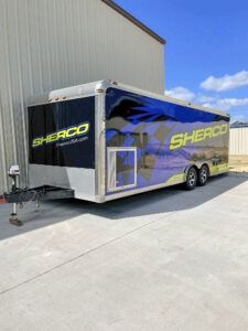 Silver Sherco enclosed trailer parked beside a building on a sunny concrete lot.