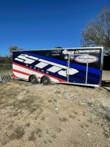 Blue, red, and white enclosed trailer parked on a dirt lot under a clear sky.