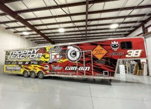 Red and yellow Can-Am Trophy Truck race vehicle in a warehouse garage, side view with sponsor graphics