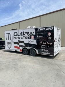 White Can-Am trailer with “Outlaw Productions” branding parked outside a warehouse under a blue sky