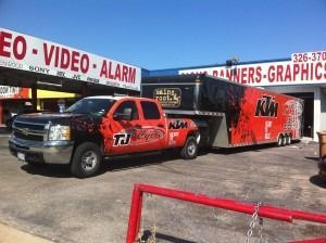 Red truck towing a black-and-red trailer parked outside a storefront.
