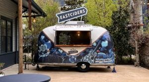 Small food trailer with “EASTCIDER” sign, open serving window, and blue mural art in a courtyard