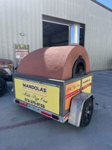 Small trailer with a brown rounded cover parked outside a warehouse, labeled Mandolas Pizza for Hire.