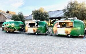 Three green food trailers parked on a paved lot near houses and trees
