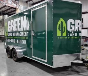 Green landscaping trailer with white lettering parked indoors, green logo on the side.