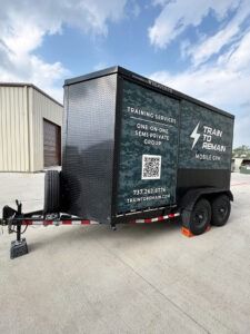 Black training trailer with QR code parked on a concrete lot beside a warehouse under a cloudy sky