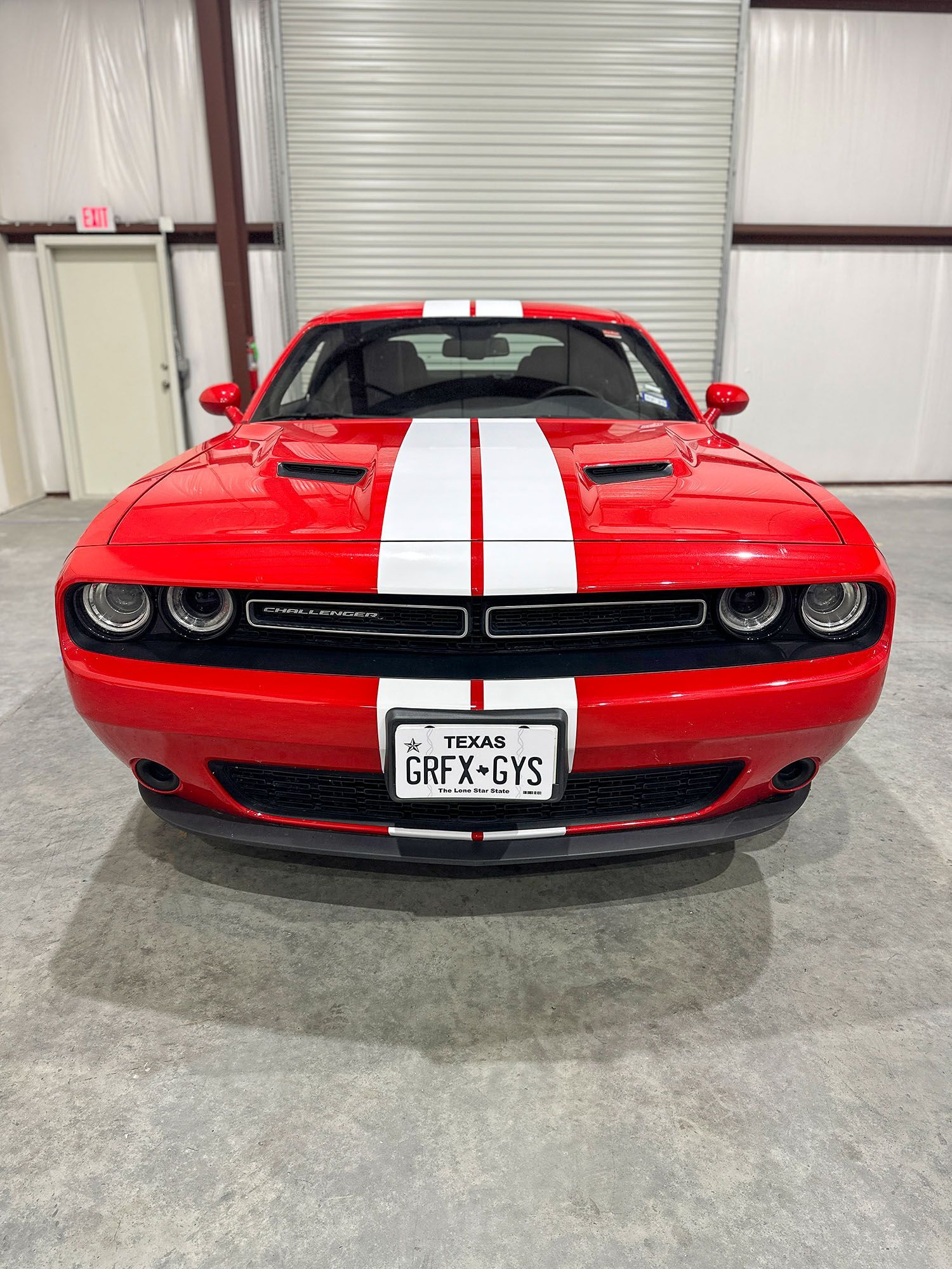 Red Dodge Challenger with white racing stripes, front view, parked indoors in a garage.