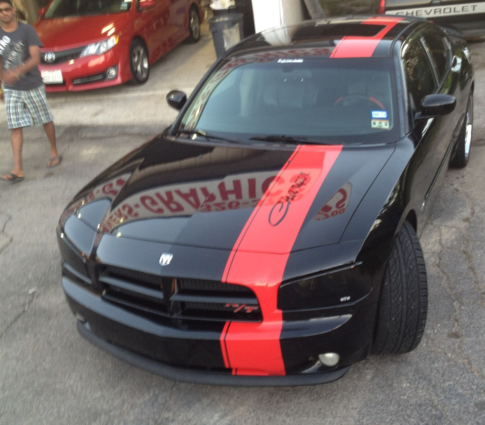 Black Dodge Charger with red racing stripe, parked at a car meet, with a person standing nearby.