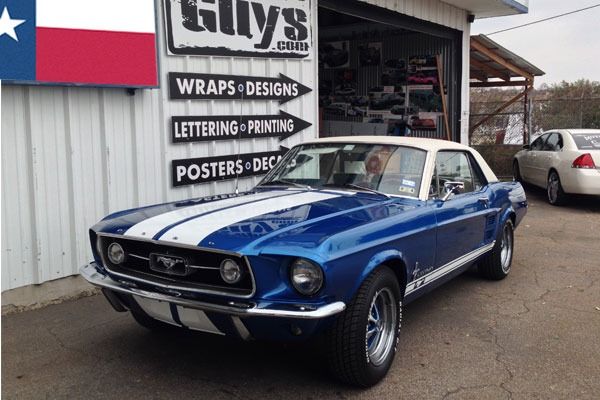Blue classic Ford Mustang parked outside a sign shop with a Texas flag mural