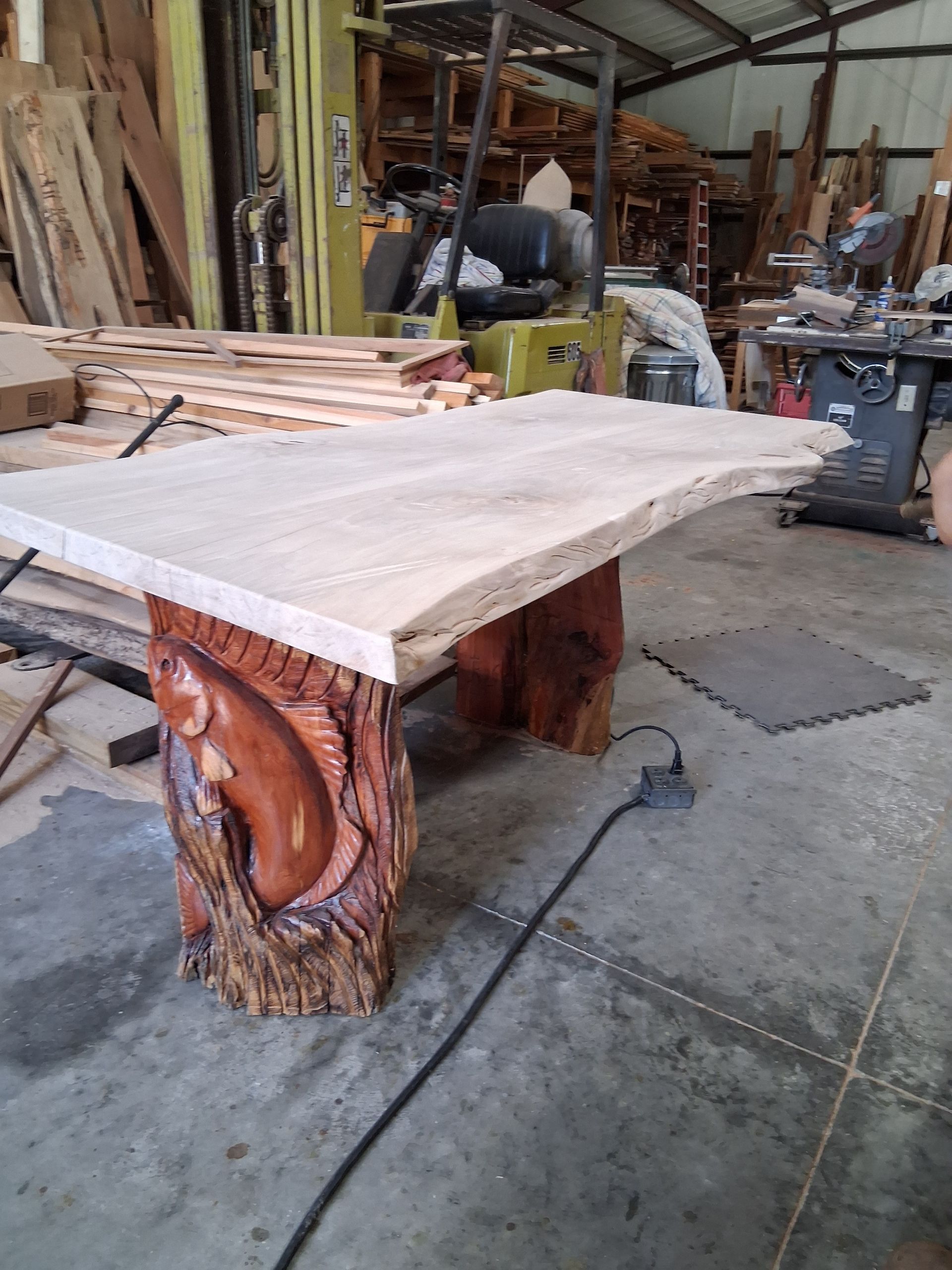 Wood slab table with carved log legs in a rustic workshop, surrounded by lumber and tools.