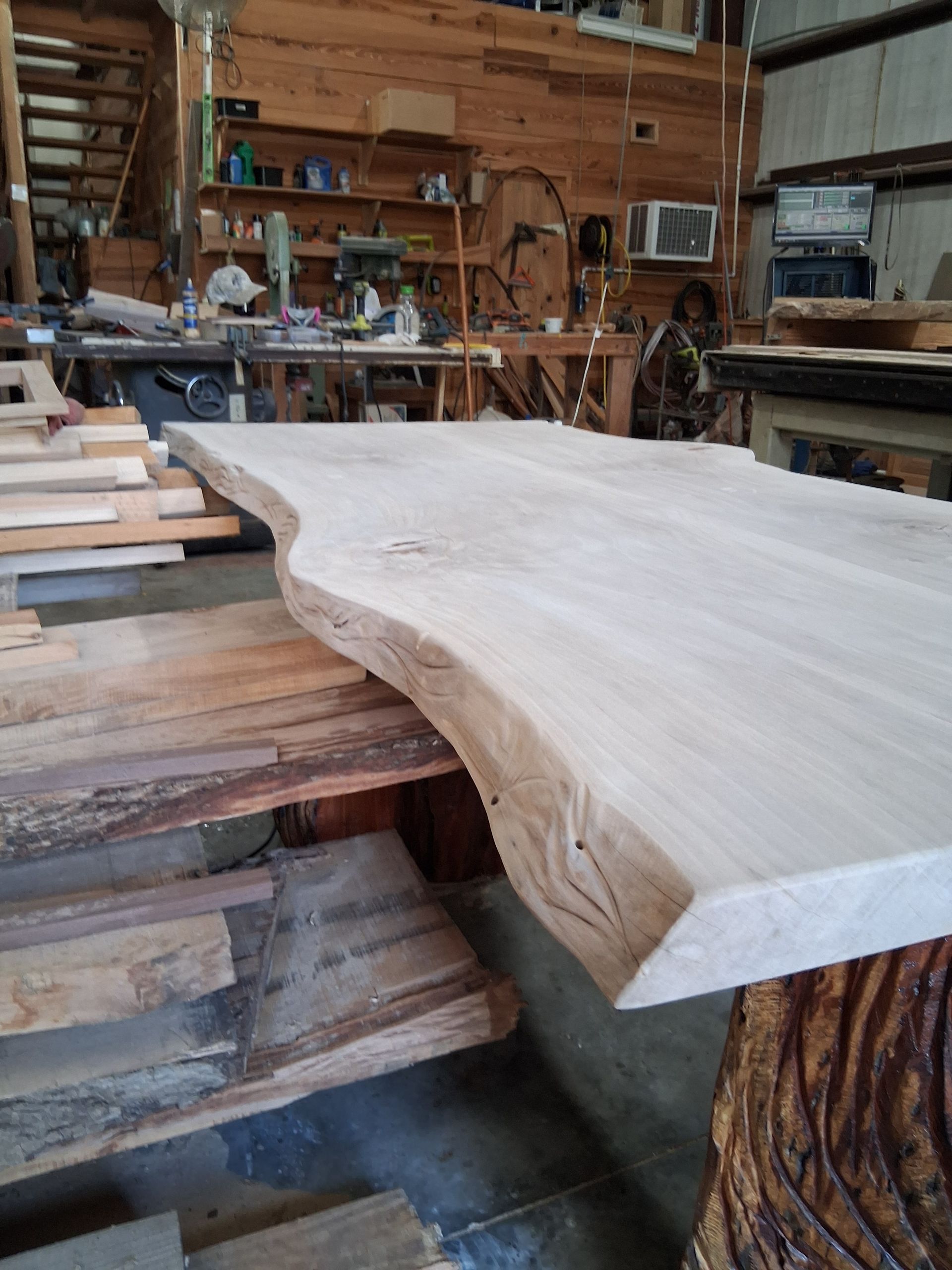 Large live-edge wood slab on a workbench in a woodworking shop, with tools and lumber in the background