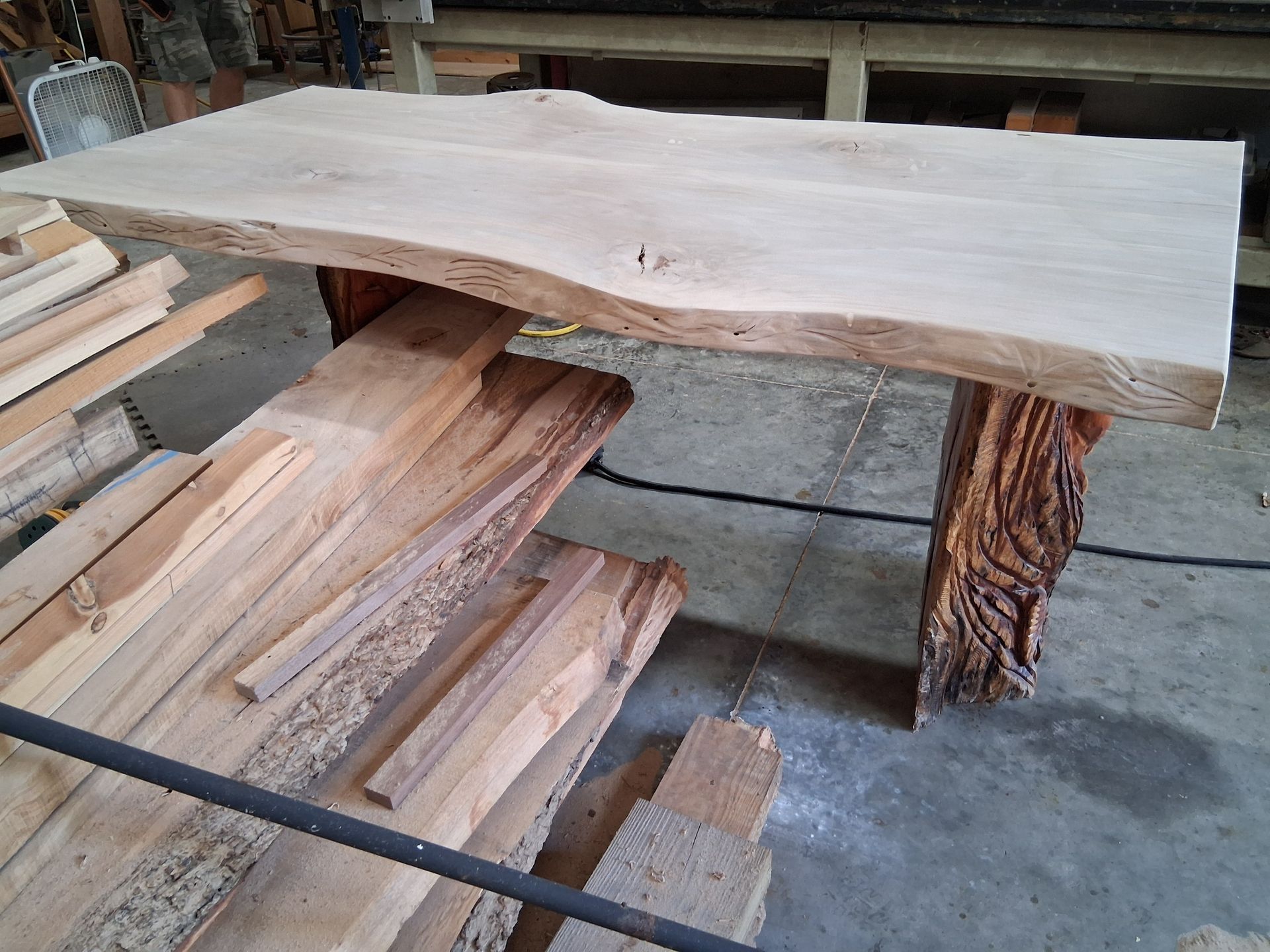 Rough live-edge wooden slab tabletop on log supports in a workshop, with stacked lumber underneath