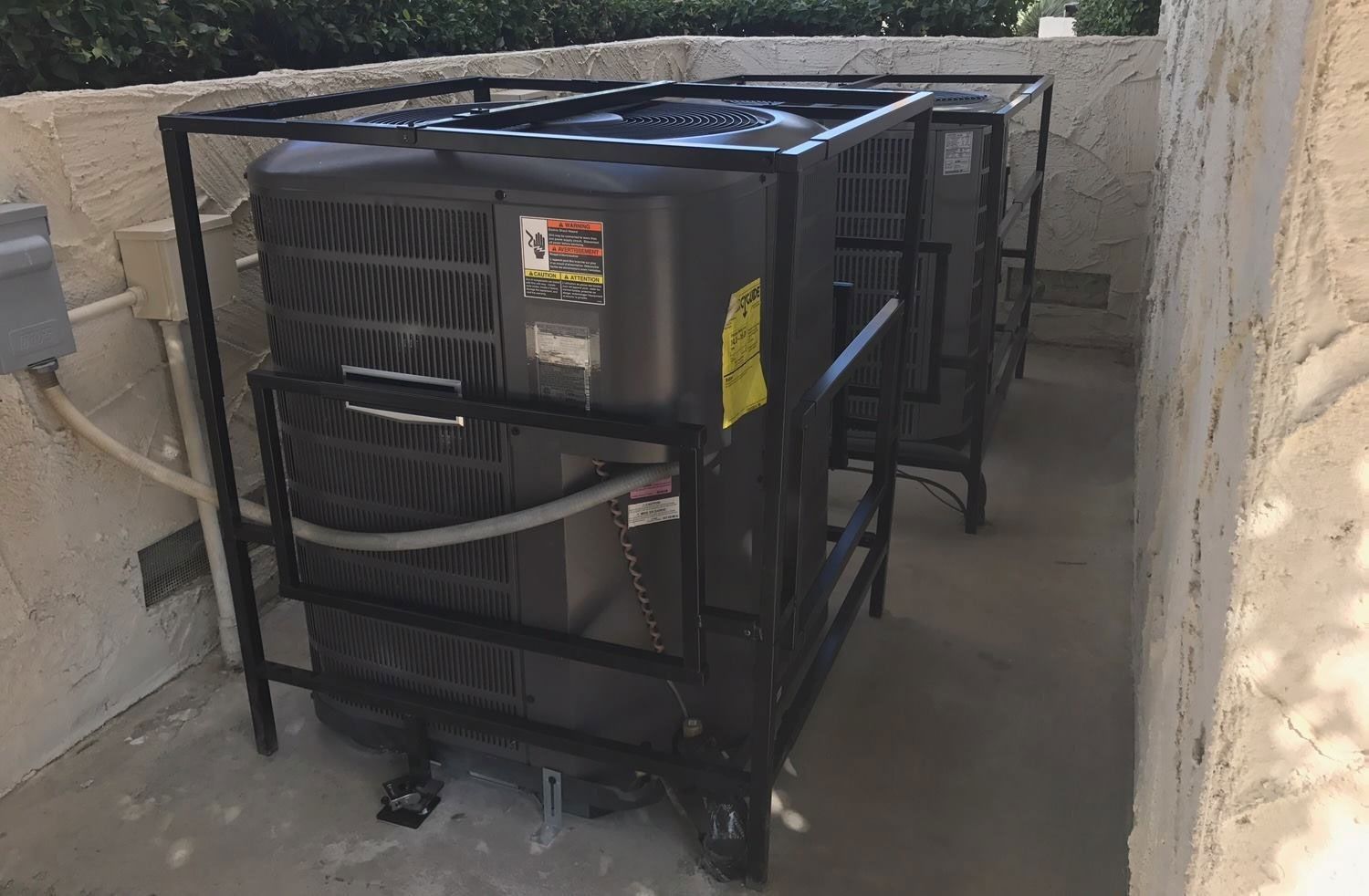 Two air conditioning units enclosed in black protective cages, located outdoors.