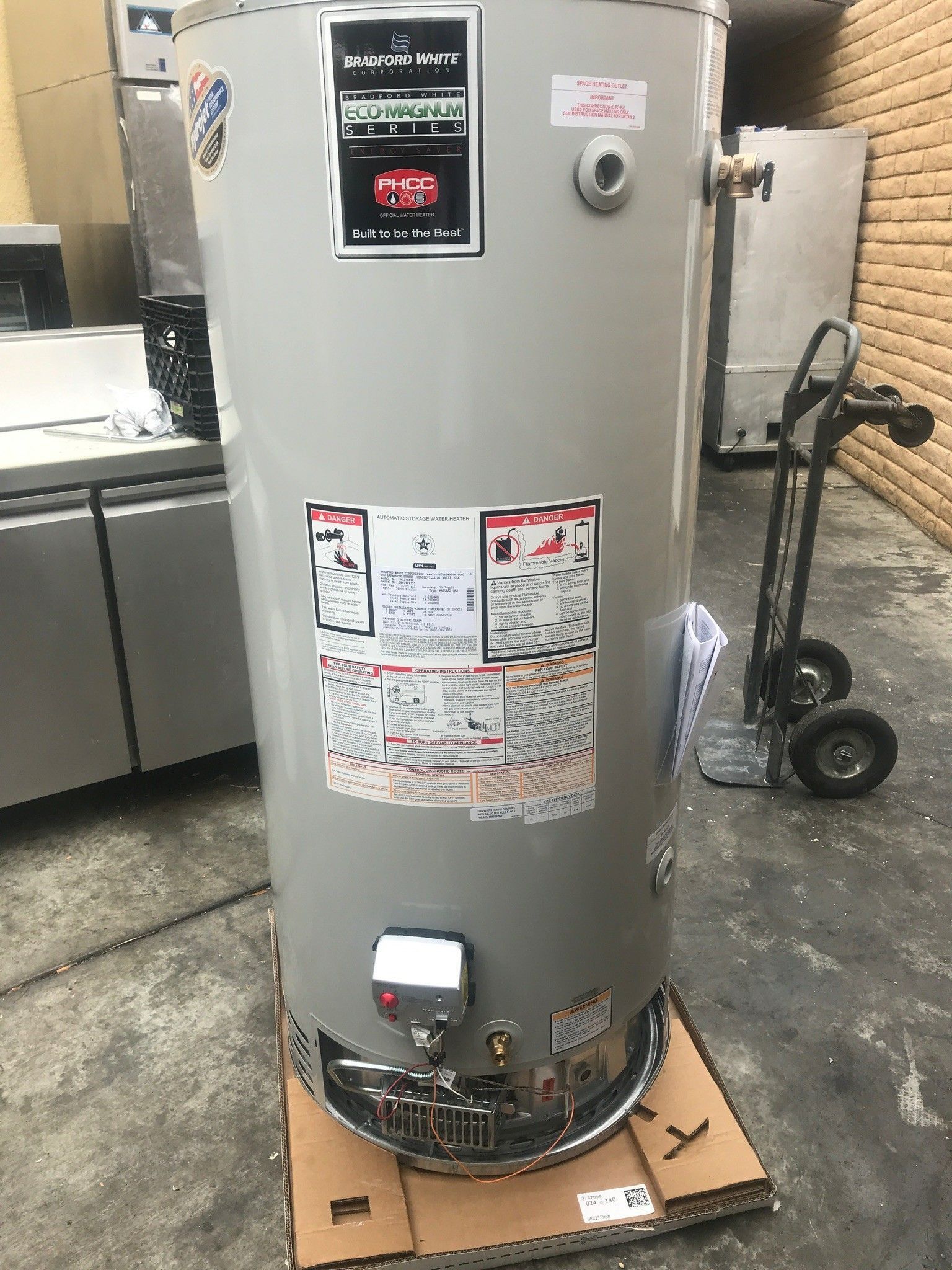 A tall, cylindrical water heater with various labels sits on a cardboard base in a warehouse setting.