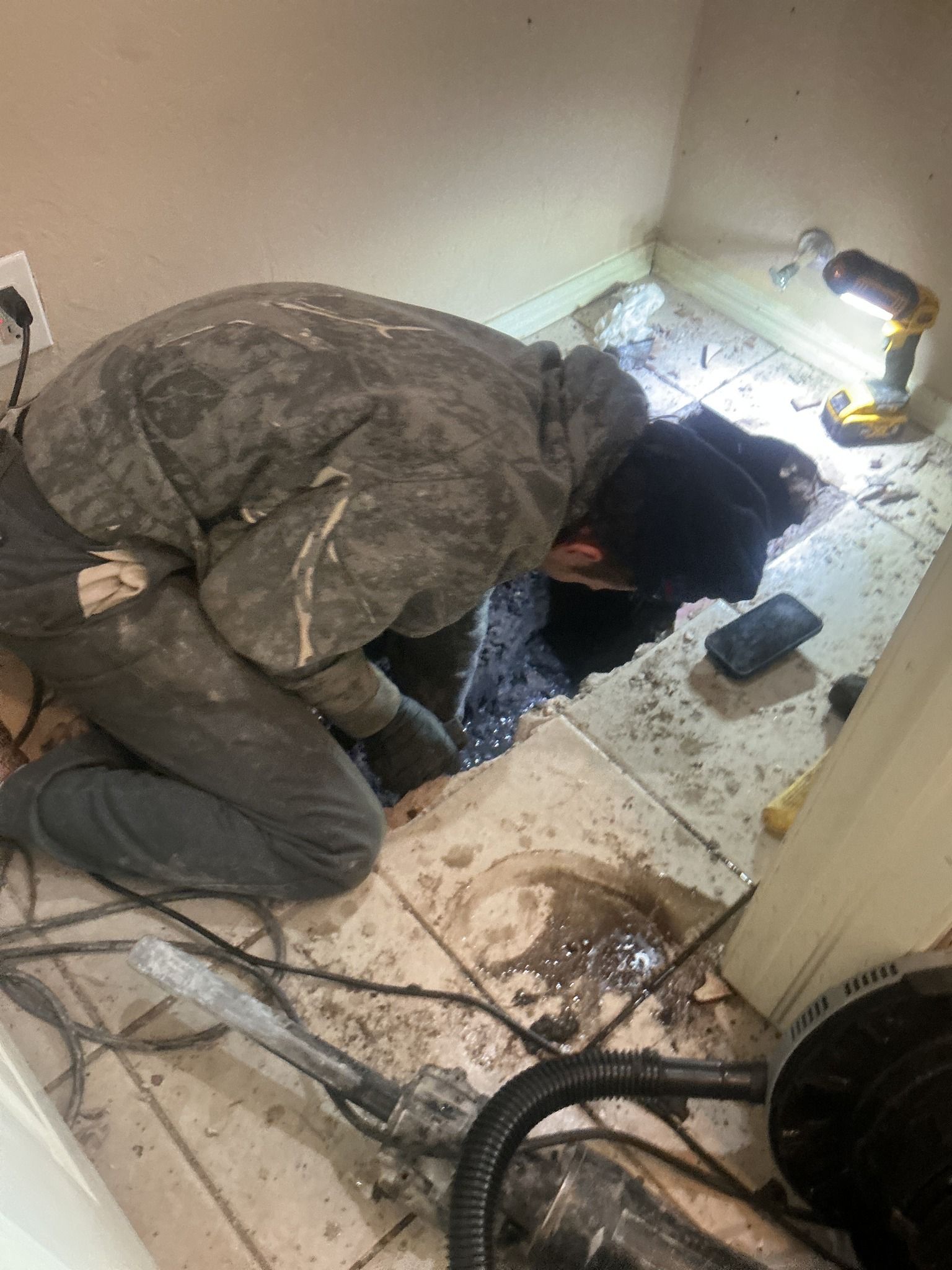 Person repairs plumbing in a room. Tile floor, tools, and a hole are visible.