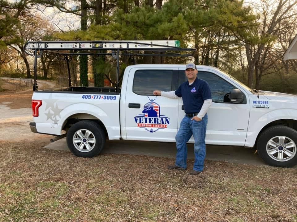 Man standing by a white truck with "Veteran" logo, ladder rack. Outdoors.