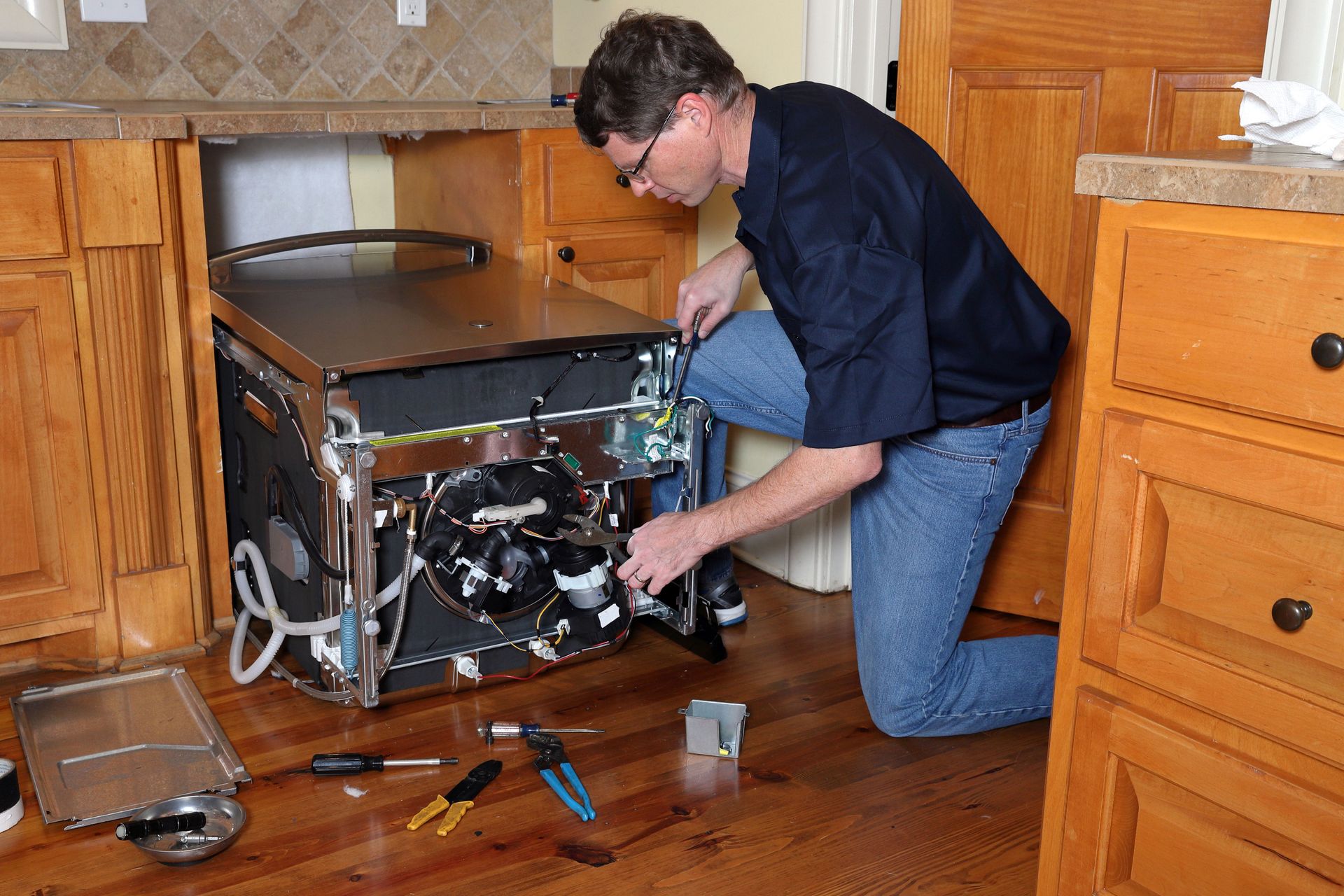 Man kneels, repairing a dishwasher in a kitchen. He wears glasses and jeans, using tools. Wooden cabinets visible.