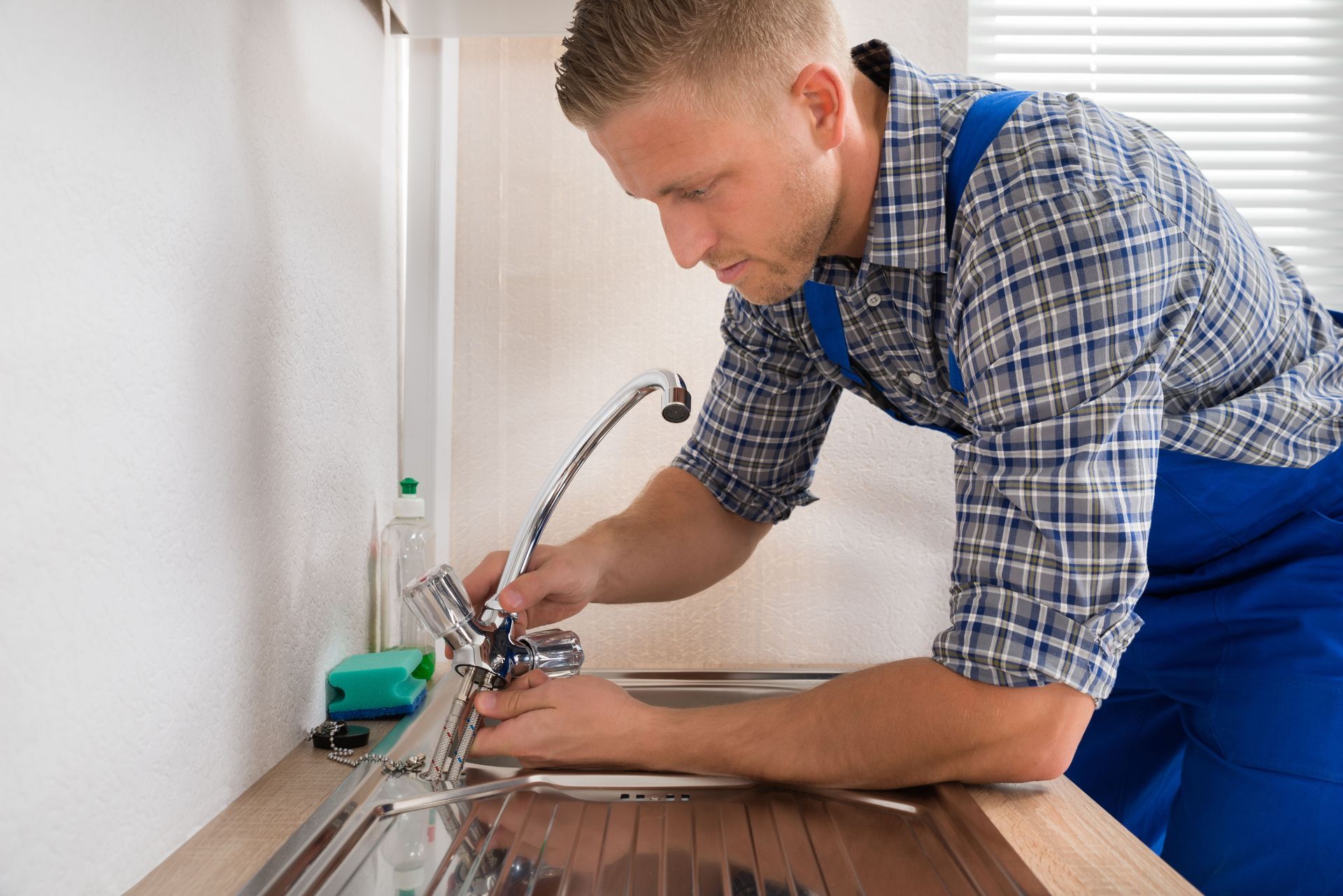 Plumber in blue overalls repairing a silver kitchen faucet at a sink.