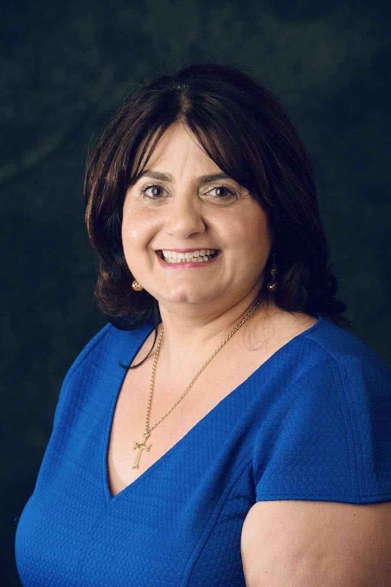 Woman with dark hair, smiling in a blue dress with a gold necklace, against a green background.