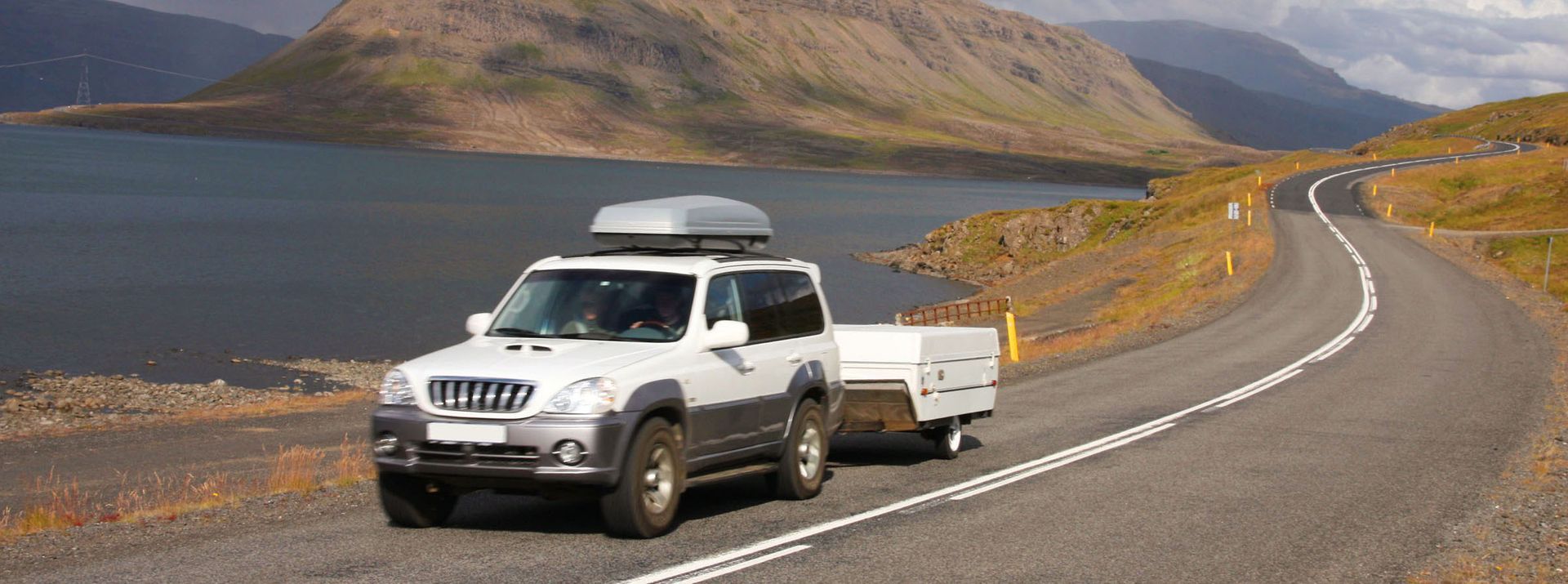 White SUV towing a small trailer on a winding road with a lake and mountains in the background.