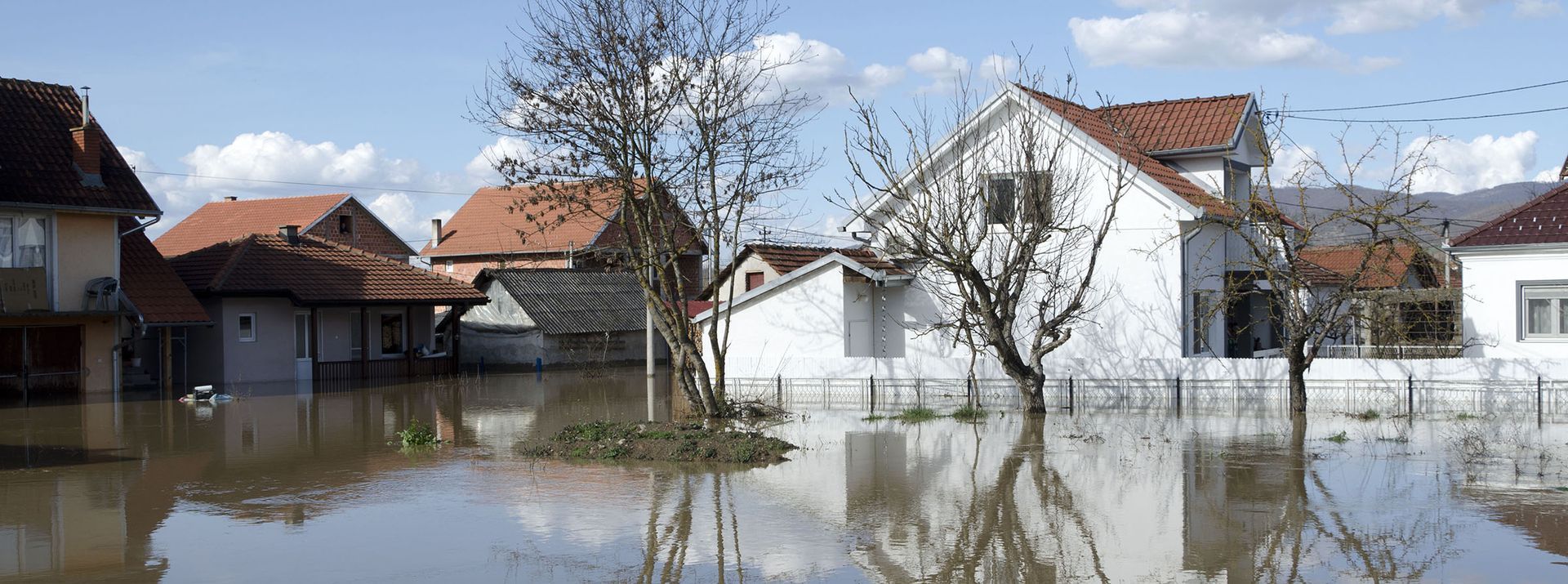 Houses flooded with brown water under a blue sky, reflecting the buildings and trees.